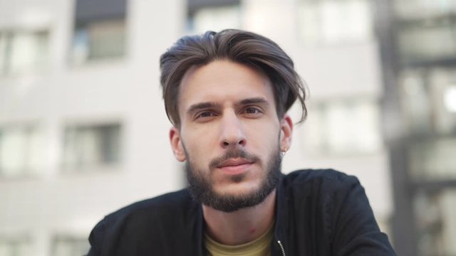 Tracking slow motion shot of handsome young man with beard and pierced ear looking attentively at camera standing outdoors in street, closeup from below view