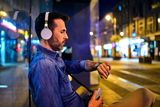 Serious man with wireless headphones waiting for night bus in the city and checking the time