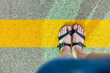 Women's feet in slippers standing at the yellow line on asphalt. Personal perspective of person looking at his feet