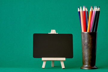 Miniature chalk board on an easel and multicolored pencils in a metal stand isolate on green background, concept back to school, selective focus