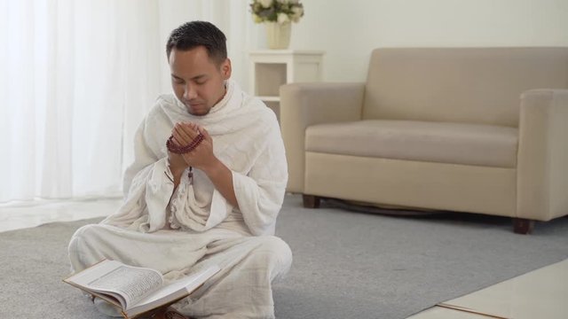 asian young man praying with Al-Qur'an and prayer beads in white traditional clothes