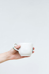 White ceramic mug in a female hand on a background of a white wall. Hand-made