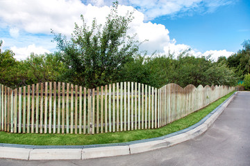 an orchard with trees growing behind a wooden fence along a corner road, an asphalt road with a curb and a drainage system on the lawn.