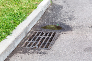 asphalt road with manhole grille at the border with green grass, drainage system with a puddle of...
