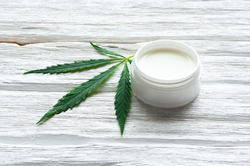 Cannabis cosmetic cream in a jar and a green plant leaf on a white wooden table background. Natural cosmetic.