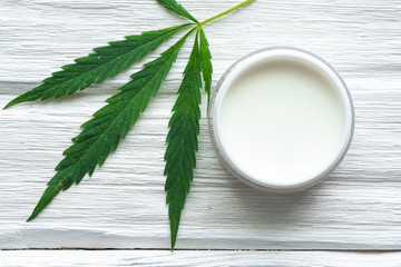 Cannabis cosmetic cream in a jar and a green plant leaf on a white wooden table background. Natural...