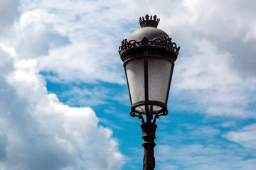 black iron street lamp with a pattern and a glass shade against the sky with clouds on a sunny day.