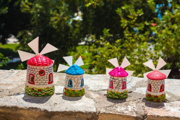 Several models of mills in bright colors. Mills stand on a stone surface against a background of green trees.