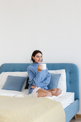 Girl in a blue sweater in interior Hygge style with a cup in hand sits on the bed