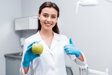 cheerful dentist holding apple and showing thumb up in clinic