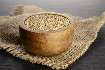 Lot of whole unpeeled sesame seeds in wooden bowl on jute cloth on grey stone