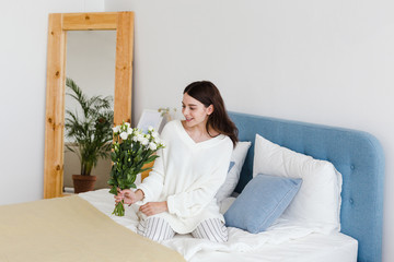Obraz na płótnie Canvas A girl in a white sweater sits on a bed holding a bouquet of white roses in her hand