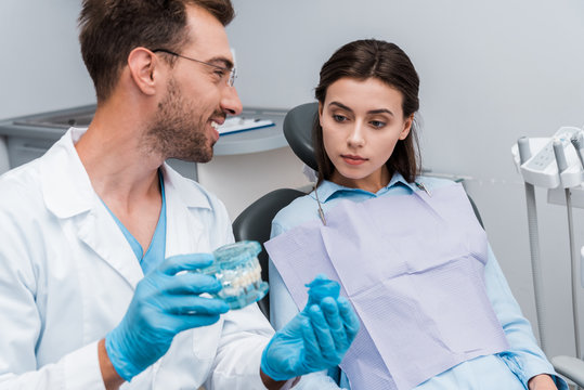 selective focus of handsome bearded man holding retainer and tooth model near attractive patient