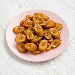 Homemade fried plantains on a pink plate on a white wooden background, side view.