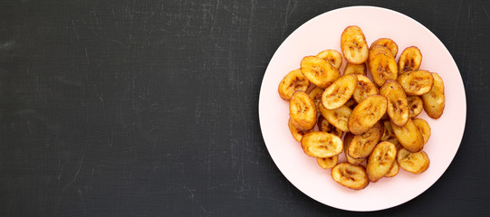 Homemade fried plantains on a pink plate on a black surface, overhead view. Flat lay, from above, top view. Space for text. - obrazy, fototapety, plakaty