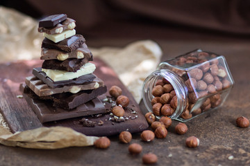 Different grades of chocolate are put the hill on a wooden board, with a filbert in a glass jar on crumpled paper, on a rusty metal surface.