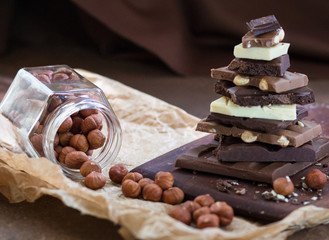 Different grades of chocolate are put the hill on a wooden board, with a filbert in a glass jar on crumpled paper, on a rusty metal surface.