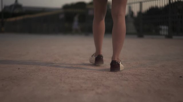 Handheld shot of slender bare legs of young woman in boots, walking along gravel path straight to gates of city park in evening time