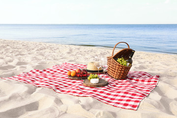 Checkered blanket with picnic basket and products on sunny beach. Space for text
