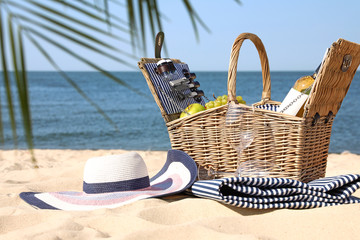 Wicker picnic basket with products, blanket and hat on sunny beach