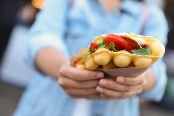 Young woman holding delicious bubble waffle with tomato and arugula outdoors, closeup