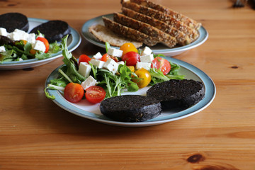 Healthy organic breakfast, two slices of black pudding with mixed leaf salad topped with yellow and red cherry tomatoes and feta cheese on a white plate with a blue rim.  Background of sliced bread.