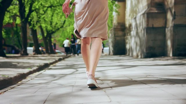 Bottom view of confident pretty woman walking along the street on lovely day. Back close-up view of feminine female legs in stylish dress and shoes. Urban city life concept.
