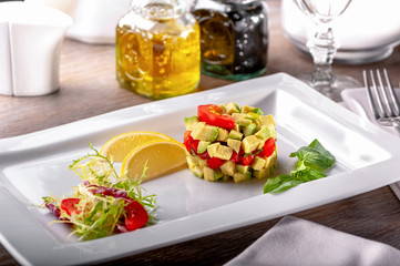 restaurant table with dish of cold appetizers tomato and avocado salad with arugula leaves, basil, lemon slices, fork on napkin, glass, gravy boats 