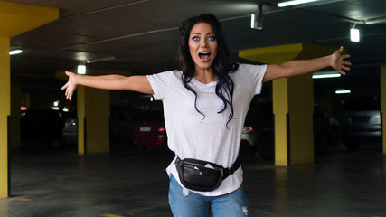 Fototapeta na wymiar Full of emotion. Waist up portrait of young woman in white t-shirt, blue jeans and fanny pack. Girl spread her arms wide. She looking at camera with wide opened eyes and raised eyebrows as if found ou