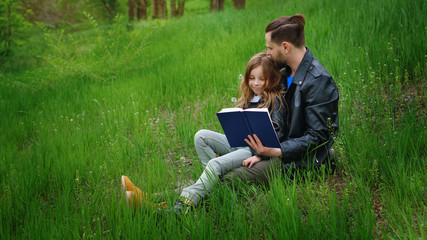 Naklejka na ściany i meble Modern stylish family walking in the park. Father and daughter are reading a book while sitting on a green lawn. Time together. Family look. Urban casual outfit