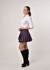 full length portrait of a brunette girl wearing a white shirt and plaid skirt uniform, standing pose in side profile against a cream studio background.