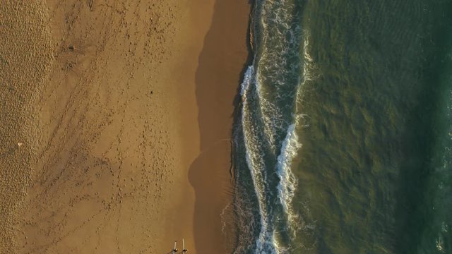 Two Surfers checking out the Waves on a beach. The Sand is lit by the orange rising sun and the surf is calm with a blueish green tone. Slowmotion