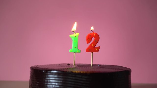 Chocolate birthday cake on pink background with green and red number twelve candle in middle electric lighter lighting candle making wish trying to blow out candle 