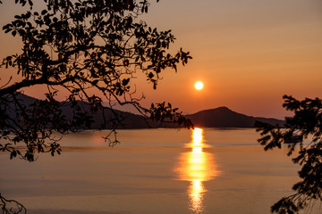 view of sunset above the mountain over the horizon on the ocean with glowing pinkish orange sky behind silhouette of tree branches with sun's reflection on the water
