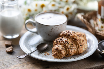 Breakfast cup of coffee and croissant with milk and peach or apricot jam on a wooden table. Bouquet of daisies. rustic