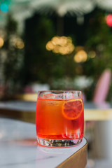 An orange aperitif cocktail in a rocks glass served on an ice cube with an orange slice in a bright colorful surrounding of a bar. Natural light. Marble table.