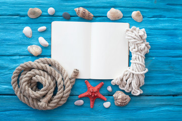 Open blank page book, red starfish, pebbles, seashells and a mooring rope on blue wooden table...