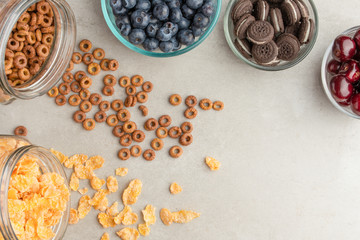 muesli and cereals are scattered with jars with berries of cherries, blueberries and sweet cookies on a light background, breakfast background