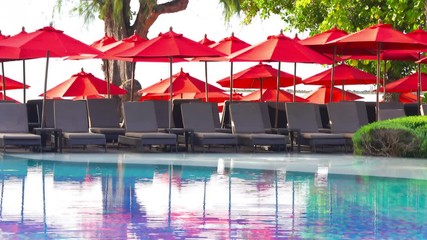 Grey sun loungers shaded by red umbrellas at the poolside of a hotel. Eye level.