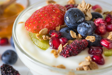 Plain yogurt with strawberry, blueberries, kiwi, granola, pomegranate in a glass bowl and honey on white wooden texture, healthy food and plant-based food concept