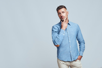 Always handsome. Confident young bearded man in blue shirt looking away and touching his chin while...