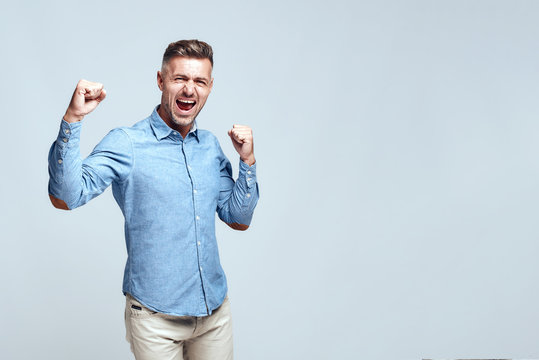Yeah, i did it Happy handsome bearded man is celebrating success and keeping hands clenched in fists while standing against grey background