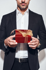Present for you. Cropped photo of handsome bearded businessman in classic suit holding a gift while standing against grey background