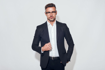 Confident look. Portrait of handsome businessman in classic suit and eyewear adjusting jacket while standing against grey background