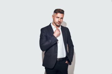 Stylish man. Bearded businessman in classic suit adjusting shirt and looking at camera while standing against grey background