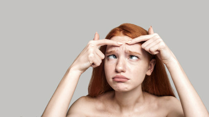 Fototapeta premium Stop acne. Worried young redhead woman examining her face while standing against grey background
