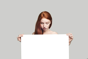 I'm intrigued. Portrait of pretty and young redhead lady holding blank sign board and looking at it...