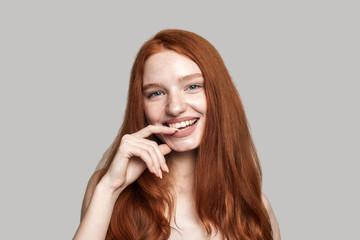 Living with smile. Studio shot of cheerful pretty young redhead lady smiling at the camera while standing against grey background
