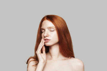 Fototapeta premium Sensuality. Portrait of beautiful woman with long red hair keeping eyes closed and touching her lips while standing against grey background