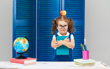Smart toddler kid. Back to school and happy time! Cute industrious child in glasses is sitting at a desk indoors. Kid is learning in class on background of blue wall. Girl reading the book.